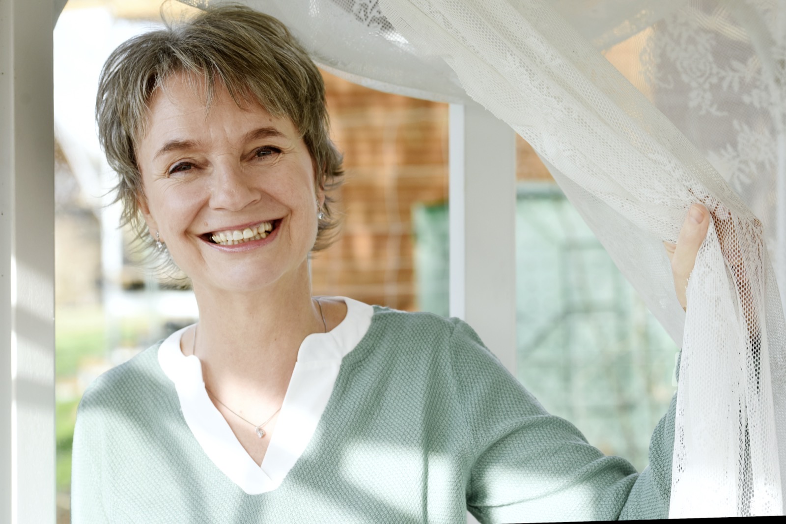 Jeanette Nentwig lacht in die Kamera und hält einen weißen Spitzenvorhang zur Seite